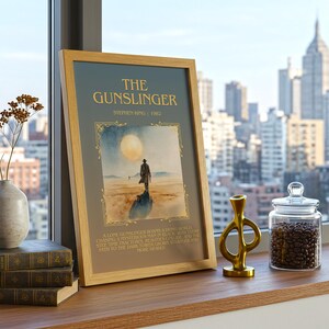 May include: A framed print of "The Gunslinger" by Stephen King, depicting a lone figure in a desert scene. Next to it are stacked books, a vase with dried flowers, a gold sculpture, and a glass jar filled with coffee beans. The scene is set by a window.