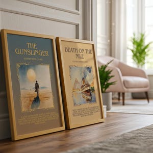 May include: Two framed book covers, "The Gunslinger" and "Death on the Nile", are propped against a wall. "The Gunslinger" shows a lone figure under a large moon. "Death on the Nile" depicts a ship. Both are in wooden frames.