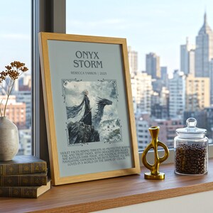 May include: A framed print titled "Onyx Storm" with a fantasy illustration of a person and a dragon. Beside it are stacked books, a vase with dried flowers, a gold sculpture, and a glass jar filled with coffee beans. The scene is set by a window with a city view.
