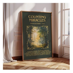 May include: A framed poster with the title "COUNTING MIRACLES" by Nicholas Sparks. The poster features a forest scene with a deer and text describing the story. The frame is gold, and the background is a dark green.