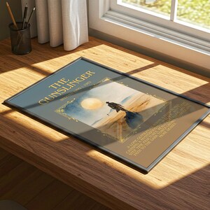 May include: Framed print of "The Gunslinger" by Stephen King, depicting a desert scene with a solitary figure. The title is in gold lettering on a dark blue background. The print rests on a wooden desk near a window.