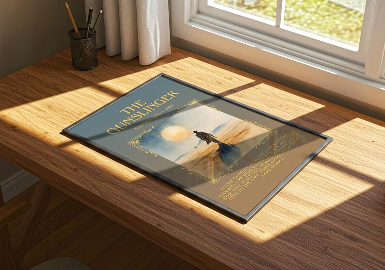 May include: Framed print of "The Gunslinger" by Stephen King, depicting a desert scene with a solitary figure. The title is in gold lettering on a dark blue background. The print rests on a wooden desk near a window.