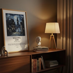 May include: A framed poster for "Salem's Lot" by Stephen King, featuring a dark, atmospheric illustration of a town street. The poster is displayed on a wooden cabinet with a lamp and bust sculptures. The lamp has a beige lampshade and wooden tripod base.