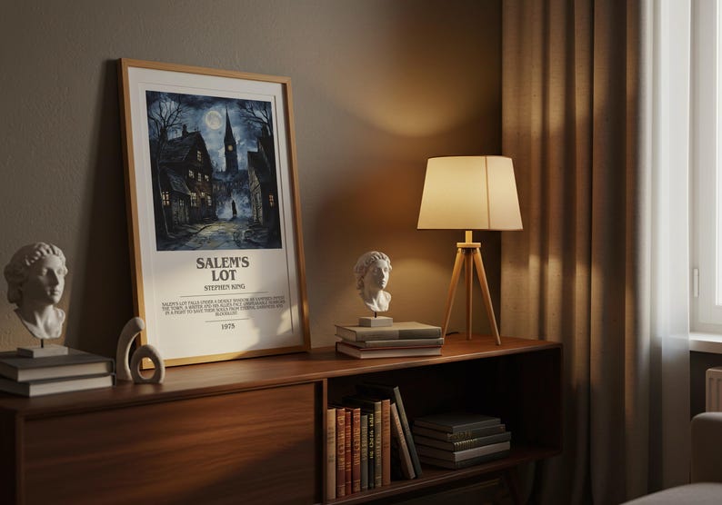 May include: A framed poster for "Salem's Lot" by Stephen King, featuring a dark, atmospheric illustration of a town street. The poster is displayed on a wooden cabinet with a lamp and bust sculptures. The lamp has a beige lampshade and wooden tripod base.