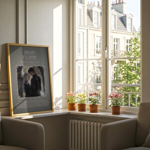 May include: A framed poster with the text "IRON FLAME" and an image of a couple. The poster is leaning against a wall next to a window with potted plants. Two armchairs are in the foreground.