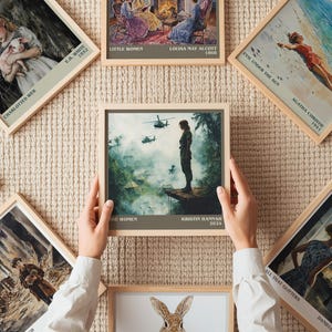May include: A collection of framed book cover art prints, including "The Women" by Kristin Hannah, "Evil Under the Sun" by Agatha Christie, and "Little Women" by Louisa May Alcott. The prints are displayed on a textured beige surface.