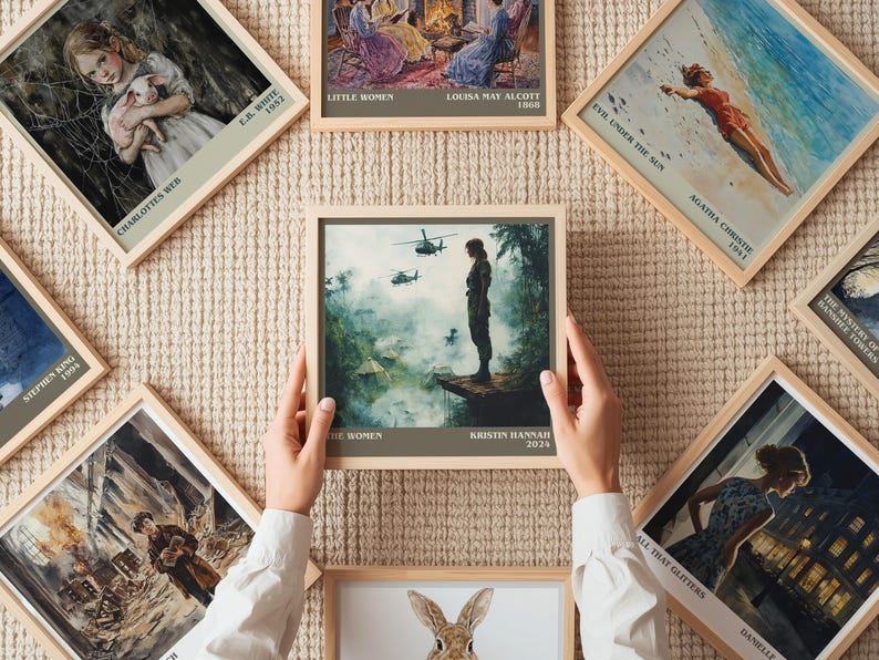 May include: A collection of framed book cover art prints, including "The Women" by Kristin Hannah, "Evil Under the Sun" by Agatha Christie, and "Little Women" by Louisa May Alcott. The prints are displayed on a textured beige surface.