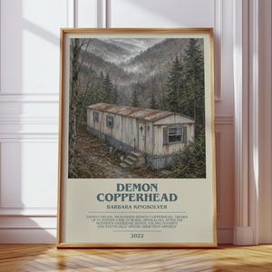 May include: A rectangular wall art print featuring a weathered mobile home in a wooded mountain landscape. Below the image, the text reads: DEMON COPPERHEAD, BARBARA KINGSOLVER, followed by a summary of the book plot and the year 2022 in dark blue.