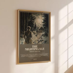 May include: A framed poster with a gold border, featuring a woman walking on a path through a forest. The poster's title is "The Nightingale" by Kristin Hannah, with text below describing the story and the year 2015.