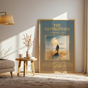 May include: A framed poster of "The Gunslinger" by Stephen King. The artwork shows a lone figure walking towards a large moon. The poster is in a gold frame and is displayed in a living room setting with a lamp and chair.