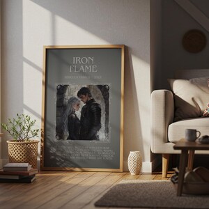May include: A framed print with the text "IRON FLAME" and an illustration of a couple. The print is in a wooden frame and is displayed in a living room setting. The room has a sofa, coffee table, and a window.