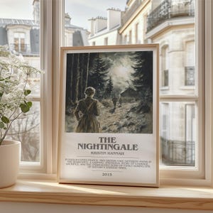 May include: A framed print of "The Nightingale" by Kristin Hannah. The artwork depicts a woman walking on a path through a forest, with text at the bottom. The frame is light-colored wood, and the print is displayed on a windowsill.