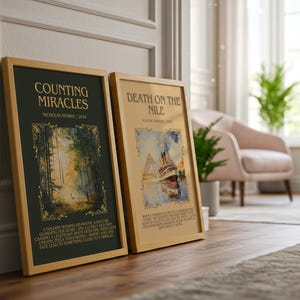 May include: Two framed book covers, "Counting Miracles" and "Death on the Nile," lean against a wall. The "Counting Miracles" cover is dark green with gold accents and forest imagery. "Death on the Nile" features a ship illustration. Both are in wooden frames.