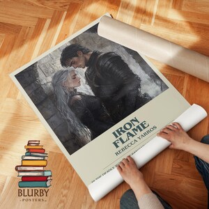 May include: A rolled poster featuring a romantic illustration of a man and woman, with the title "Iron Flame" and author Rebecca Yarros. The poster has a light beige background and is being unrolled. A stack of books is visible.