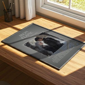 May include: A framed print titled "Iron Flame" featuring a romantic illustration of a couple. The artwork is in a dark color palette with text around the central image. The frame is black, and the print is displayed on a wooden surface.