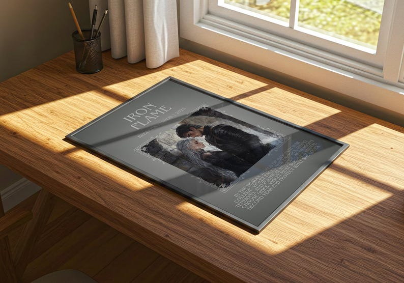 May include: A framed print titled "Iron Flame" featuring a romantic illustration of a couple. The artwork is in a dark color palette with text around the central image. The frame is black, and the print is displayed on a wooden surface.