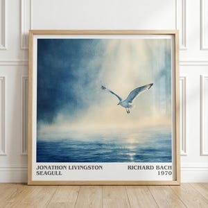 May include: Framed art print featuring a seagull in flight over water. The artwork has a soft, watercolor-like quality with shades of blue and white. Text at the bottom reads "JONATHON LIVINGSTON SEAGULL" and "RICHARD BACH 1970". The frame is made of light-colored wood.