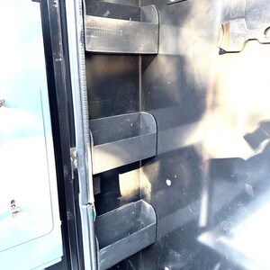 May include: A close-up of a black metal storage compartment with three black plastic shelves. The shelves are mounted on the side of the compartment and are designed to hold small items.