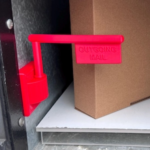 May include: A bright red, 3D-printed "Outgoing Mail" sign attached to a metal surface. The sign is positioned next to a brown cardboard box, suggesting a mail drop-off location. The sign's text is clearly visible.