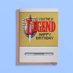 May include: A rectangular greeting card featuring pixel art of a silver sword and a blue and gold shield. The text reads YOU'RE A LEGEND HAPPY BIRTHDAY in red and black pixelated font. The card has a gold centre panel and white borders.