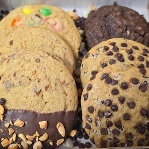 May include: A box of assorted cookies, including chocolate chip, peanut butter, and M&M cookies. The cookies are arranged in a box with a brown paper liner. The box is filled with peanuts.