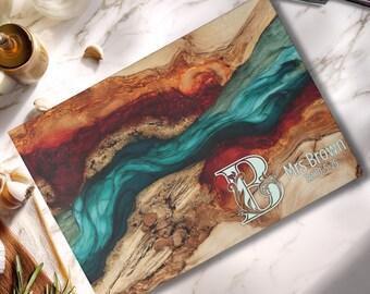 May include: A rectangular wooden chopping board with a teal and red resin river design. The board is inscribed with "Mrs Brown" and the date "5-5-25", plus a decorative initial "B". A chef's knife is on the marble surface.