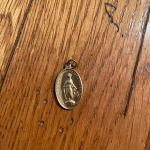 May include: A gold-colored metal pendant featuring a depiction of the Virgin Mary. The pendant has an inscription in French that reads "Mère du Bon Conseil, priez pour nous".