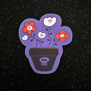 May include: A purple pot with a skull face holds white and red flowers with smiling faces. The flowers have green stems and leaves.