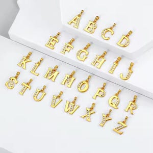 Puede incluir: Una colección de colgantes de letras iniciales doradas, cada uno con un pequeño lazo para una cadena. Se muestran las letras de la A a la Z, algunas con pequeñas piedras preciosas transparentes. Los colgantes están dispuestos sobre una superficie blanca.