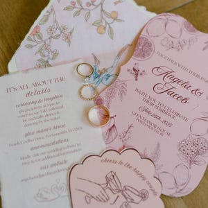 Op de afbeelding: Een verzameling trouwbriefpapier, waaronder uitnodigingen, RSVP-kaarten en een decoratief label. De uitnodigingen zijn roze met bloemen- en citrusillustraties. Er zijn ook gouden trouwringen aanwezig. De tekst op de uitnodiging luidt "Angela & Jacob" en de datum "25 juli 2025."
