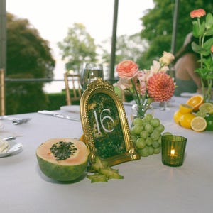 K&ouml;nnte beinhalten: Eine Tischdekoration mit einer goldgerahmten Tischnummer 16, frischem Obst, Blumen und einer brennenden Kerze. Der Tisch ist f&uuml;r eine Veranstaltung gedeckt, mit einer wei&szlig;en Tischdecke und Glaswaren.