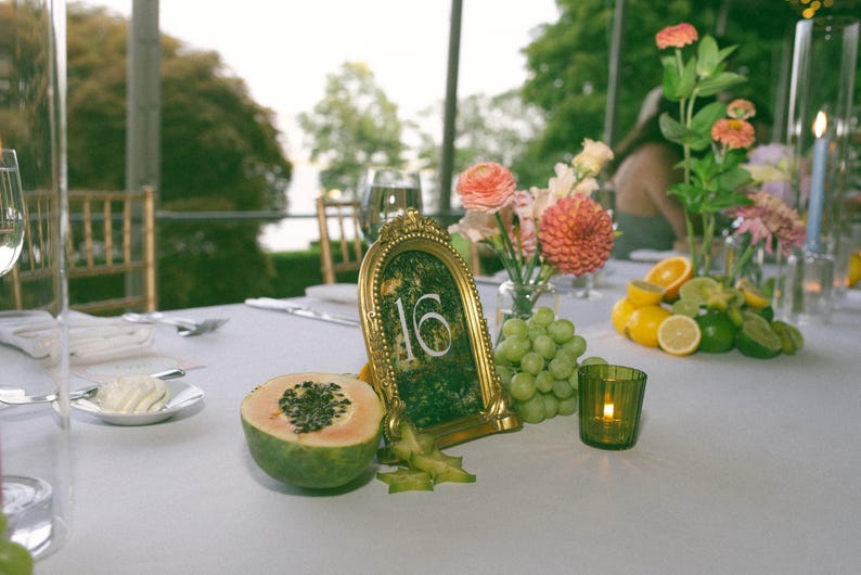 K&ouml;nnte beinhalten: Eine Tischdekoration mit einer goldgerahmten Tischnummer 16, frischem Obst, Blumen und einer brennenden Kerze. Der Tisch ist f&uuml;r eine Veranstaltung gedeckt, mit einer wei&szlig;en Tischdecke und Glaswaren.