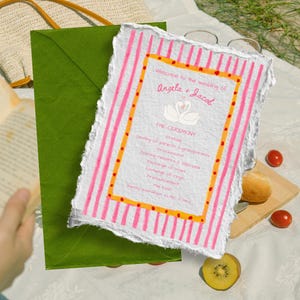 Könnte beinhalten: Eine Hochzeitseinladung mit rosa und weißen Streifen, mit den Namen "Angela + Jacob" und einer Schwan-Illustration. Die Einladung befindet sich auf strukturiertem Papier, mit einem grünen Umschlag im Hintergrund.