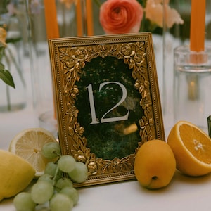 Puede incluir: Un número de mesa con marco dorado con el número 12, rodeado de detalles florales. La mesa está adornada con fruta fresca como limones, uvas y naranjas. Velas naranjas altas y arreglos florales en el fondo.