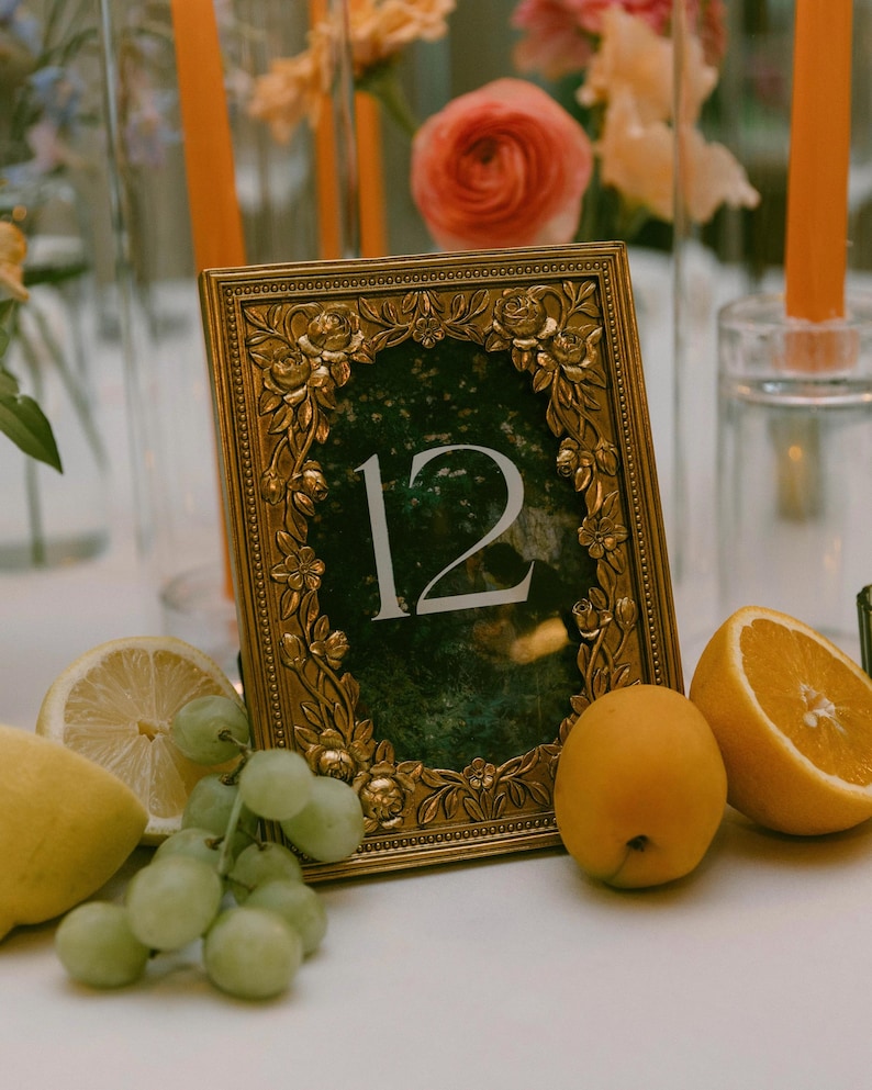 K&ouml;nnte beinhalten: Ein goldgerahmter Tischaufsteller mit der Zahl 12, umgeben von floralen Details. Die Tischdekoration umfasst frische Fr&uuml;chte wie Zitronen, Trauben und Orangen. Hohe orangefarbene Kerzen und Blumenarrangements sind im Hintergrund.