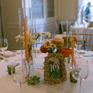 K&ouml;nnte beinhalten: Eine Hochzeitstafel mit Weingl&auml;sern, Blumengestecken und orangefarbenen Kerzen in Glasvasen. In der Mitte befindet sich eine goldgerahmte Tischnummer 10, mit einer Traube gr&uuml;ner Trauben. Das Tischtuch ist wei&szlig;.