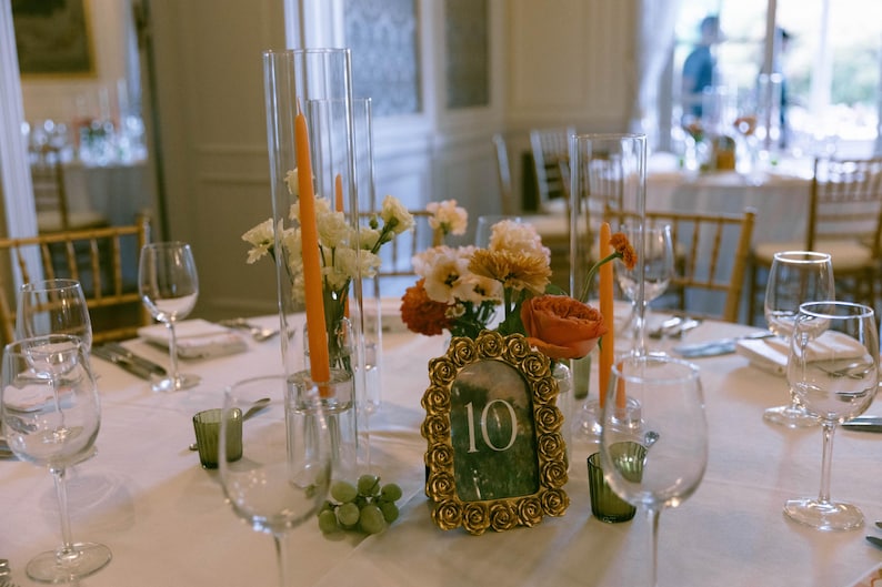 K&ouml;nnte beinhalten: Eine Hochzeitstafel mit Weingl&auml;sern, Blumengestecken und orangefarbenen Kerzen in Glasvasen. In der Mitte befindet sich eine goldgerahmte Tischnummer 10, mit einer Traube gr&uuml;ner Trauben. Das Tischtuch ist wei&szlig;.