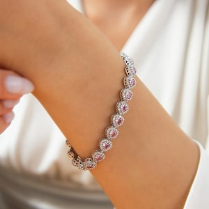 May include: A silver bracelet featuring heart-shaped pink gemstones, each surrounded by smaller, clear stones. The bracelet is worn on a wrist, showcasing its delicate design and sparkle. The jewelry is set against a white background.