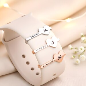 May include: A cream-coloured smartwatch strap adorned with three personalised heart-shaped charms. The charms are silver and rose gold, each bearing a letter and a date. One charm reads "Diana", another "J", and the third "X".