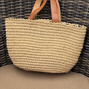 May include: A woven straw tote bag with brown leather handles. The bag is a light tan color and has a rounded shape. The handles are attached to the top of the bag and are made of a smooth, light brown leather.