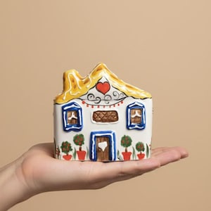 Può includere: Un oggetto decorativo a forma di casa in ceramica bianca, con tetto giallo e dettaglio a cuore rosso. La casa ha finestre e porta con cornice blu e piante in vaso alla base. Tenuto in una mano.