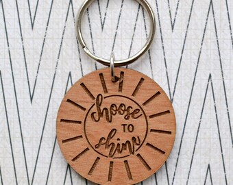 May include: Wooden keychain with a sun design and the text "choose to shine" engraved on it.