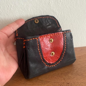 Könnte beinhalten: Eine kleine schwarze Ledermünzbörse mit einem roten Lederklappen und orangefarbenen Nähten. Die Börse hat zwei Messing-Druckknöpfe auf der Klappe.