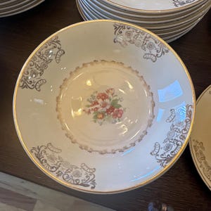 May include: A white ceramic bowl with a gold rim and floral design. The bowl has a scalloped edge and a floral pattern in the center. The bowl is on a wooden surface.