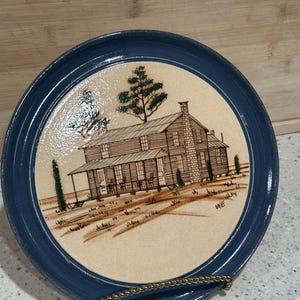 May include: A ceramic plate featuring a hand-painted rural cabin scene with a blue border. The cabin has a stone chimney and a porch. The plate is displayed on a gold-coloured metal stand.