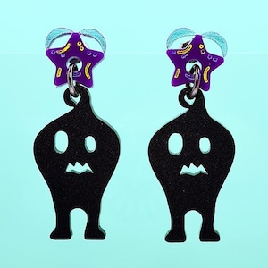 Puede incluir: Un par de pendientes con dijes negros en forma de fantasma con acentos en forma de estrella púrpura y amarilla.