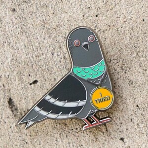 May include: An enamel pin of a pigeon wearing a green collar and a yellow medal that says "I TRIED".