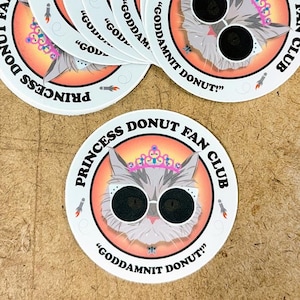 May include: A set of six round white stickers with a pink and white cat wearing sunglasses and a crown. The cat is surrounded by a pink and orange circle with the text "Princess Donut Fan Club" and "Goddamnit Donut!" in black text.
