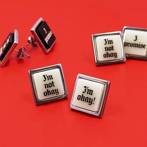 May include: A collection of square stud earrings with silver-toned metal frames. Some feature black centers with white text, while others have off-white centers with black text. The text reads I'm not okay, I'm okay!, and I promise in a gothic font.