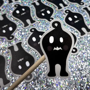 Puede incluir: Pegatinas negras y blancas con forma de fantasma con un diseño de dibujos animados. Cada pegatina presenta una cara sonriente con ojos blancos y una boca pequeña. Las pegatinas están sobre un fondo plateado brillante, y una está unida a un palito de madera.
